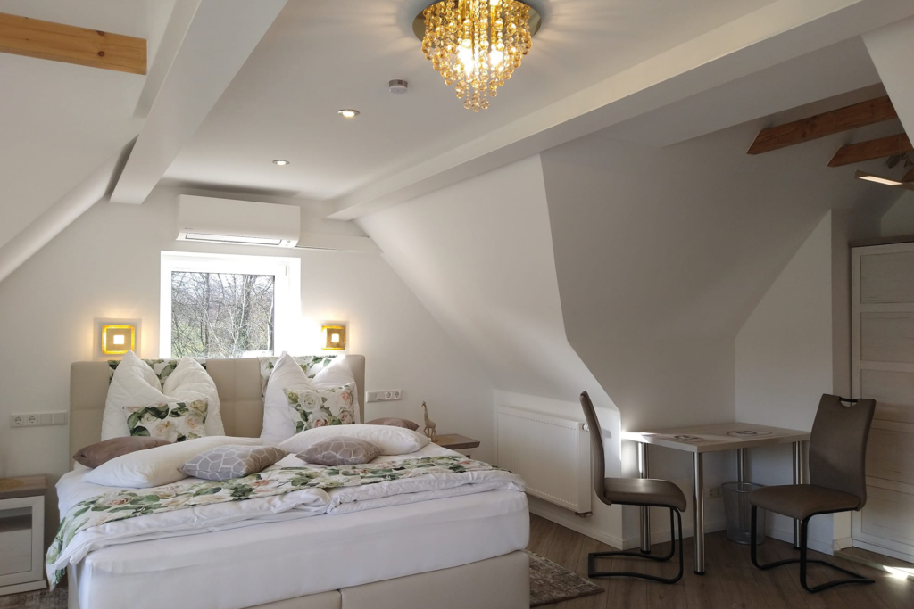 Helles Dachzimmer mit Doppelbett, floraler Bettwäsche, Kristallleuchter und kleinem Esstisch mit zwei Stühlen.