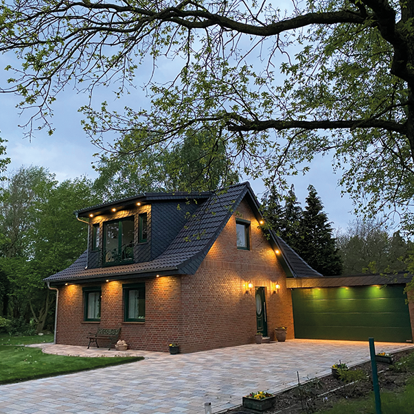 Modernes Einfamilienhaus aus rotem Klinker mit beleuchteter Fassade und grünem Garagentor in abendlicher Stimmung.