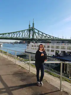 Frau steht lächelnd am Flussufer vor einer Brücke und einem vorbeifahrenden Schiff.