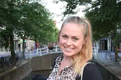 Frau mit blondem Haar lächelt in die Kamera auf einer Stadtstraße mit Bäumen im Hintergrund.