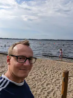 Mann mit Brille macht ein Selfie am Strand mit Blick aufs Wasser.