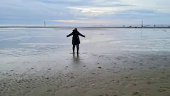 Person steht mit ausgebreiteten Armen am nassen Strand bei Ebbe.