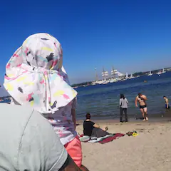Kind mit Sonnenhut am Strand blickt aufs Meer, im Hintergrund Segelschiffe und Badegäste.