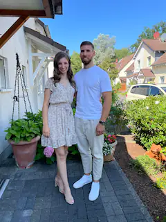 Junges Paar steht lächelnd im Garten vor einem Haus bei sonnigem Wetter.