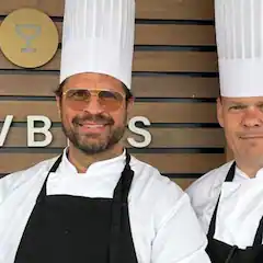 Zwei Köche in weißer Uniform und Kochmütze lächeln in die Kamera.