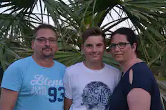 Familie mit Sohn steht vor grünen Palmen und lächelt in die Kamera.