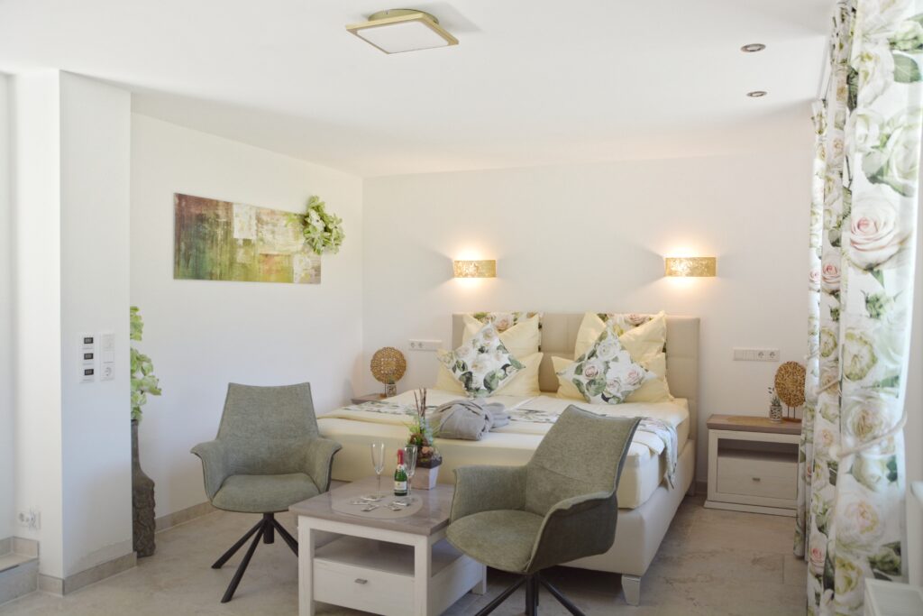 Helles Schlafzimmer mit Doppelbett, floraler Bettwäsche und zwei grünen Sesseln.