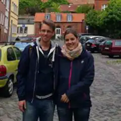 Zwei Personen stehen lächelnd auf einer Kopfsteinpflasterstraße vor geparkten Autos und alten Häusern.